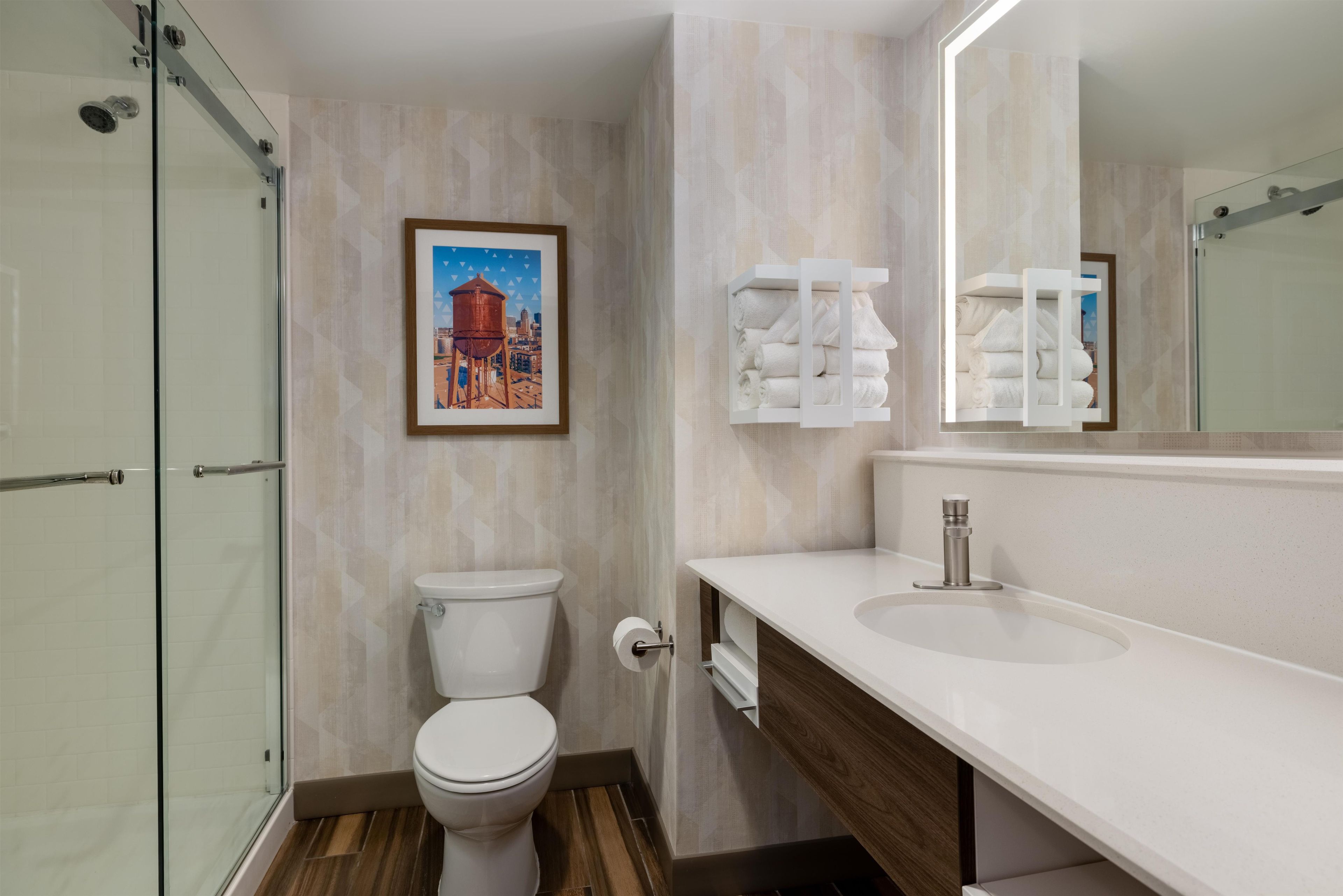 Baño con cabina de ducha de vidrio, inodoro, lavabo con espejo y un cuadro enmarcado en la pared.