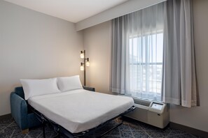 Une chambre d’hôtel avec un lit, un fauteuil bleu, une fenêtre avec des rideaux et un climatiseur fixé au mur.