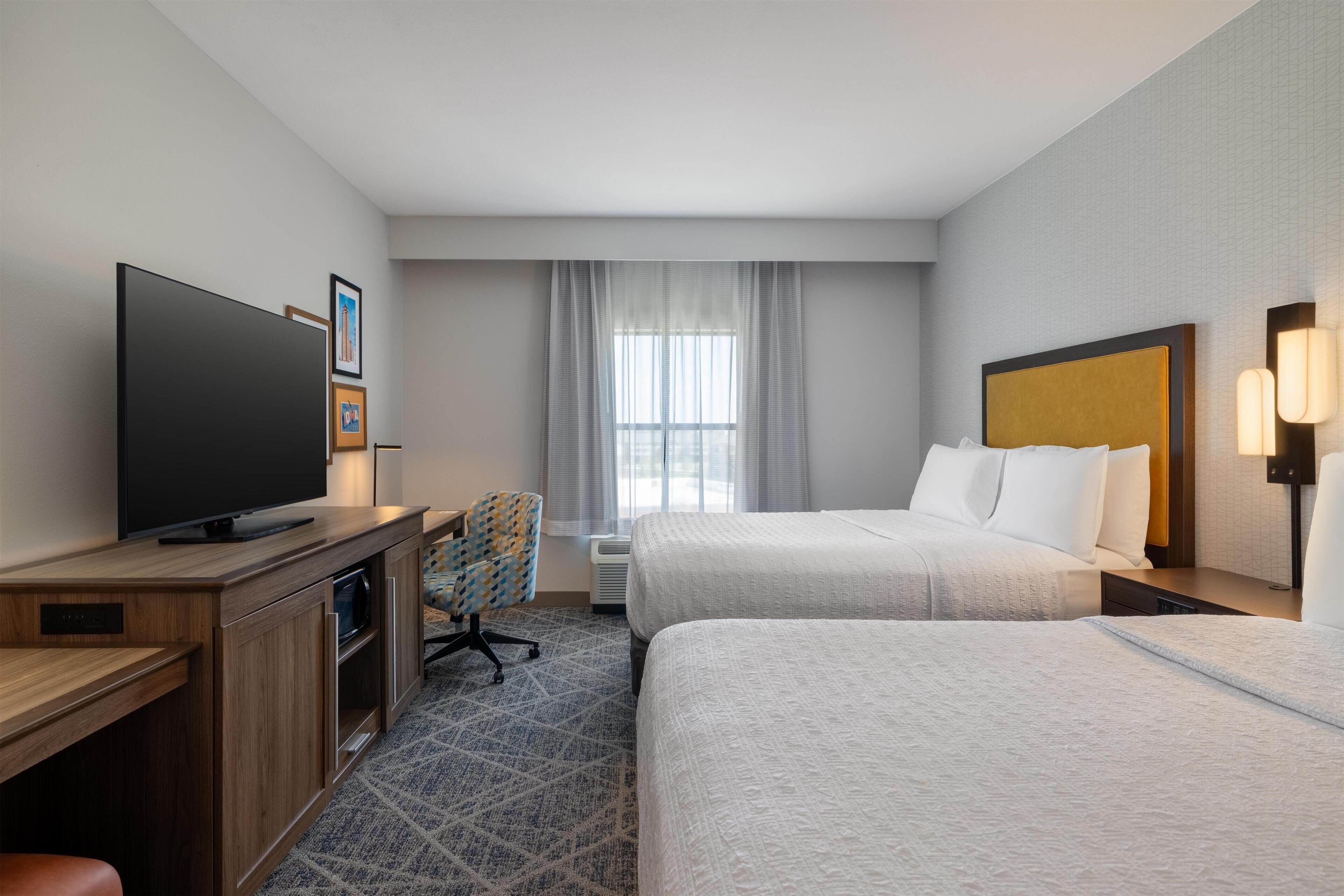 Habitación de hotel con dos camas, un escritorio con televisor, una silla y una ventana con cortinas.