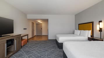 A hotel room with a flat-screen TV, a wooden desk, two beds, a patterned carpet, and a hallway leading to a door.