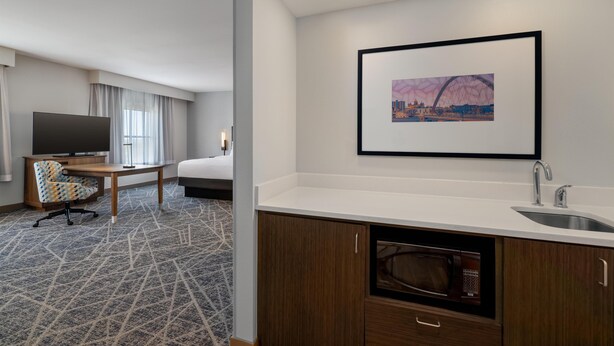 Ein Hotelzimmer mit Bett, Schreibtisch mit Stuhl, Fernseher, einem gerahmten Bild an der Wand, einem Waschbecken und einer Mikrowelle.