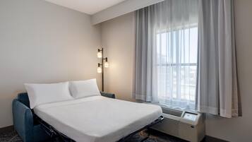 A hotel room with a bed, a blue armchair, a window with curtains, and a wall-mounted air conditioning unit.