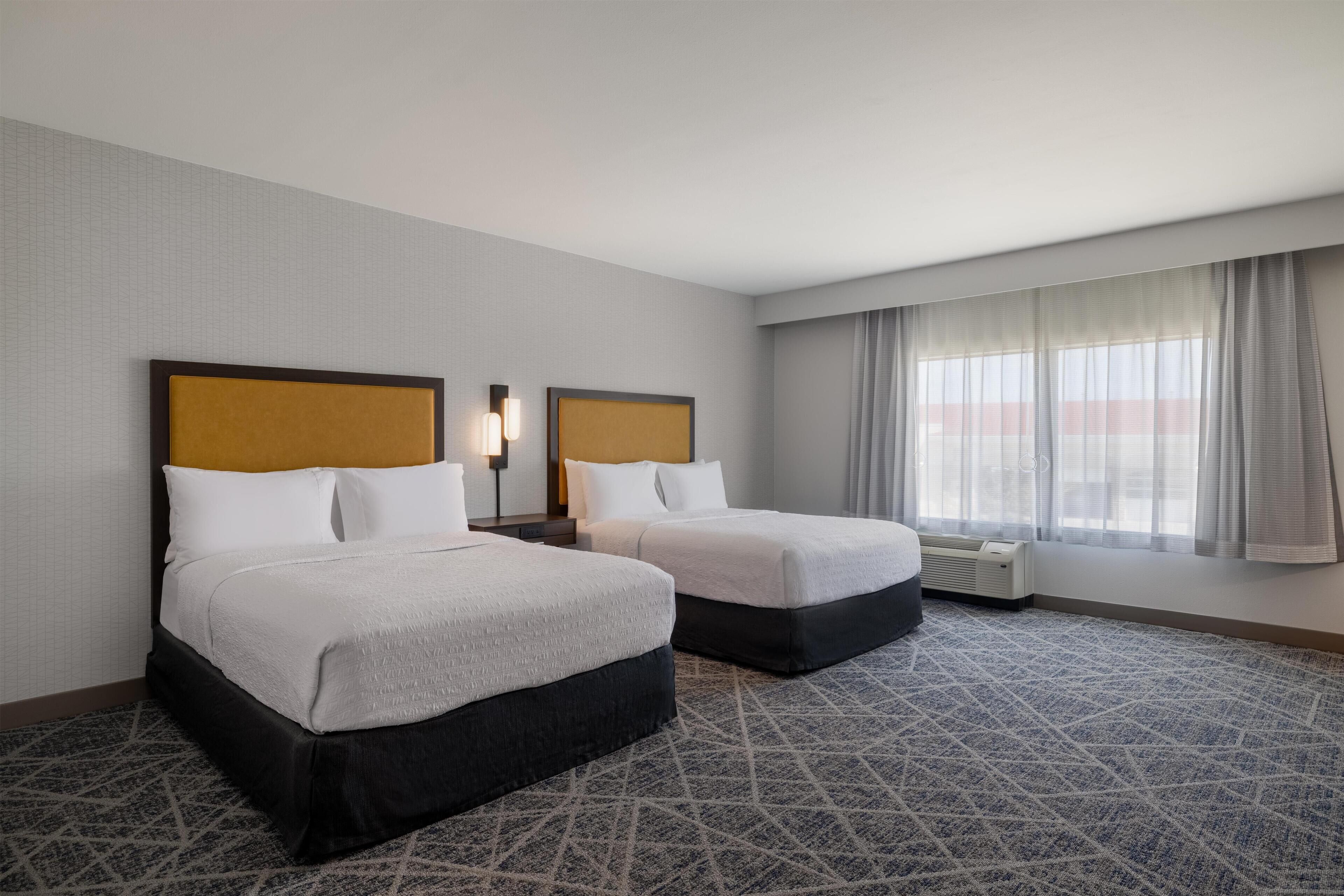 A hotel room with two beds, a large window with curtains, a patterned carpet, and a wall-mounted air conditioning unit.