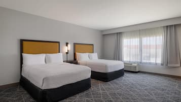 A hotel room with two beds, a large window with curtains, a patterned carpet, and a wall-mounted air conditioning unit.