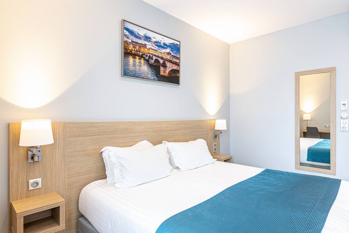 Habitación de hotel con una cama grande, dos lámparas de noche, una mesita de noche, un cuadro colgado en la pared y un espejo.