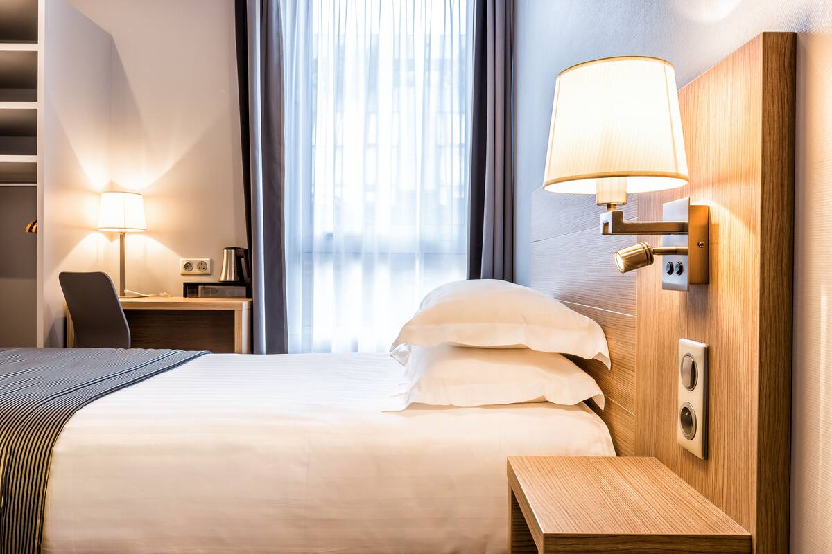 Habitación de hotel con cama, mesita de noche, lámpara, escritorio, silla y vistas al exterior a través de amplias ventanas.