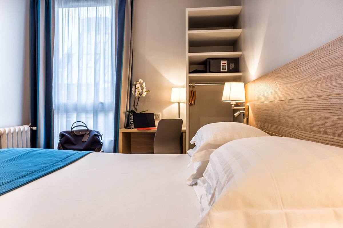 Habitación de hotel con cama, escritorio con una computadora portátil, silla, lámpara, estante con una caja fuerte y un bolso en el suelo.