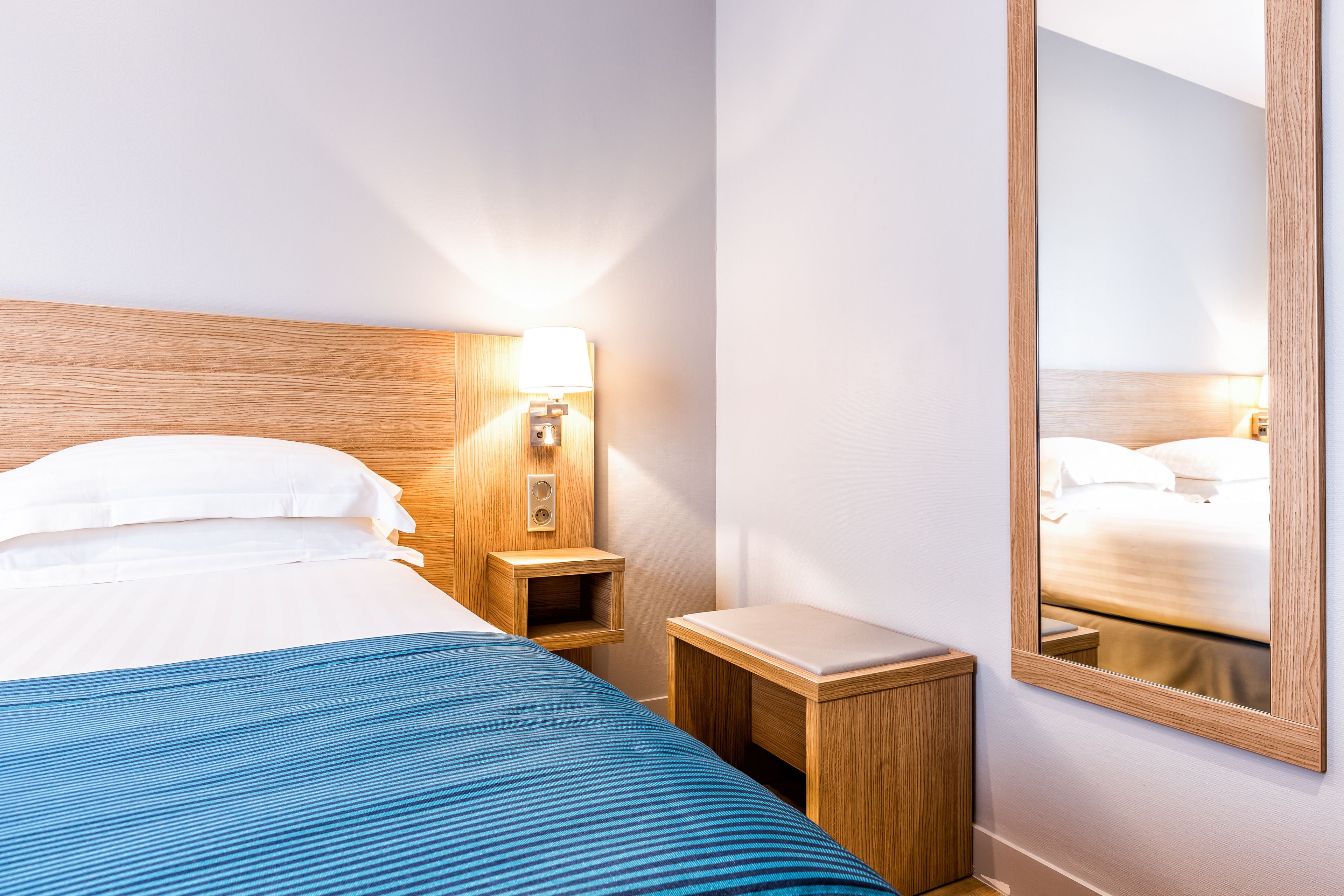 A hotel room with a bed, a bedside table with a lamp, a small wooden nightstand, and a mirror on the wall.