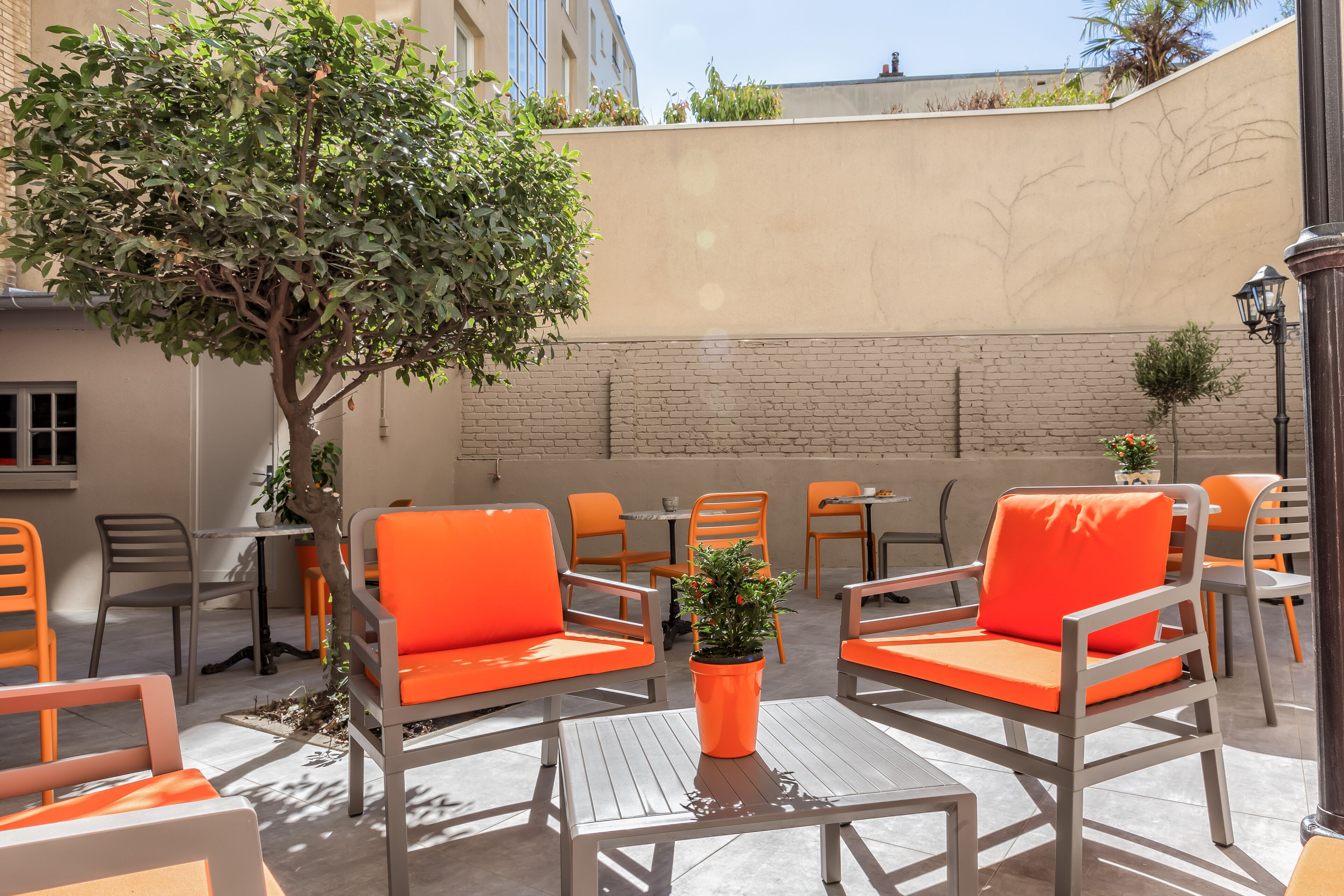 An outdoor seating area with orange chairs and tables, a tree, and a brick wall.