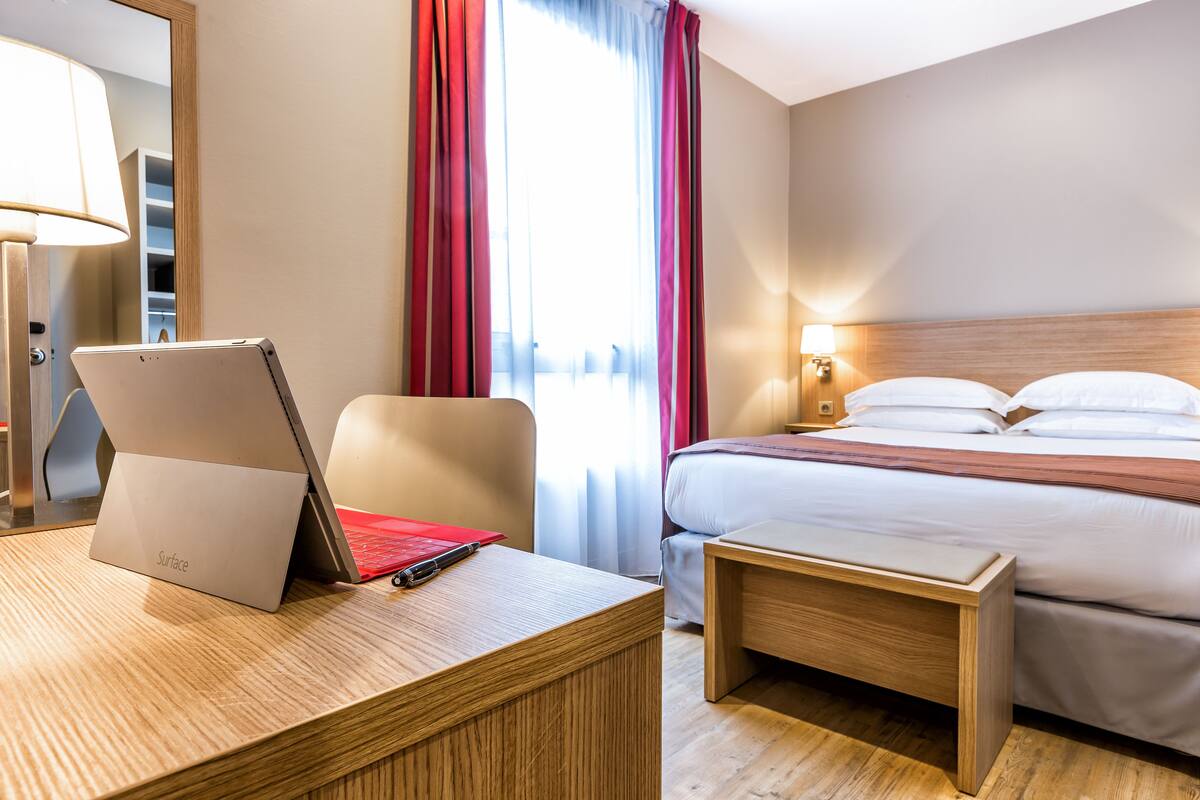 Una habitación de hotel con cama, mesita de noche, lámpara y un ordenador portátil sobre el escritorio.