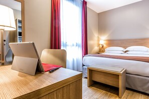 Ein Hotelzimmer mit Bett, Nachttisch, Lampe und einem Laptop auf dem Schreibtisch.