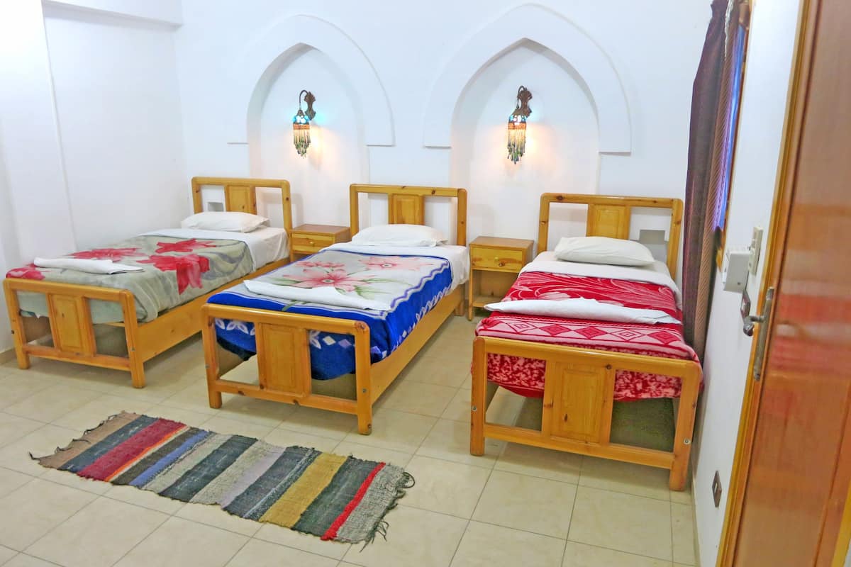 Una habitación con tres camas de madera, cada una con un edredón estampado diferente. Hay dos lámparas de pared sobre las camas, y una alfombra a rayas en el suelo.