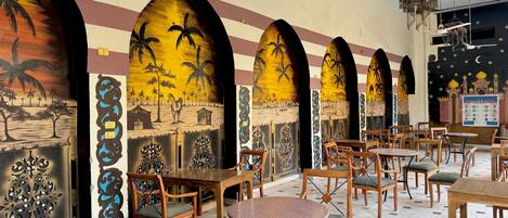 L’intérieur d’un restaurant, avec de nombreuses tables et chaises, des peintures murales décoratives et un sol carrelé.