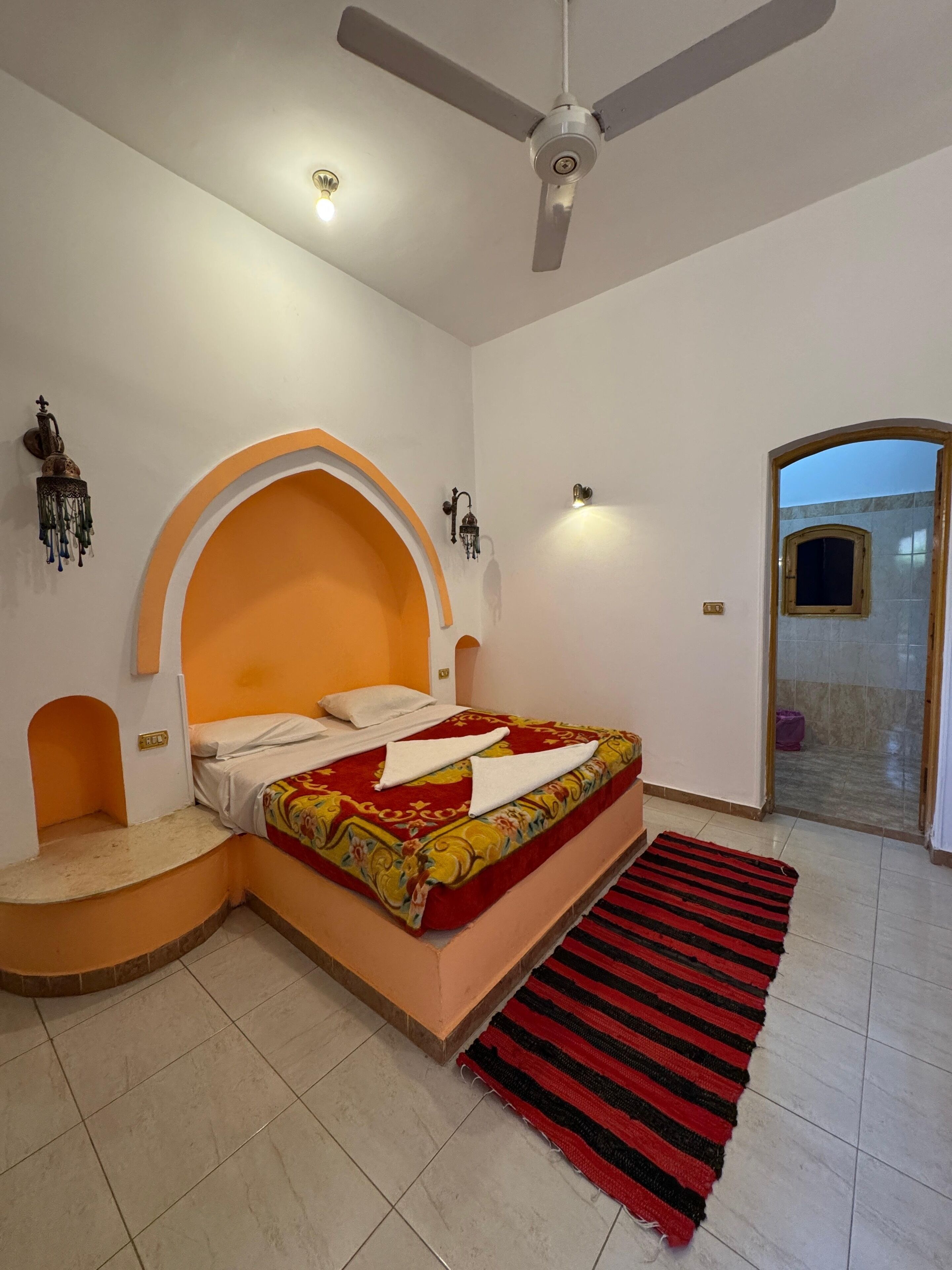 Een slaapkamer met een bed, een plafondventilator, een zwart-rood gestreept tapijt en een deur naar een badkamer.
