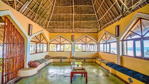 A room with a thatched roof, large windows, and a central seating area with cushions.