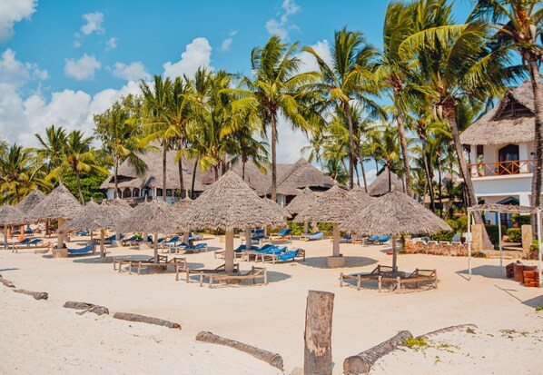 Een strand met rieten parasols, palmbomen en ligstoelen.