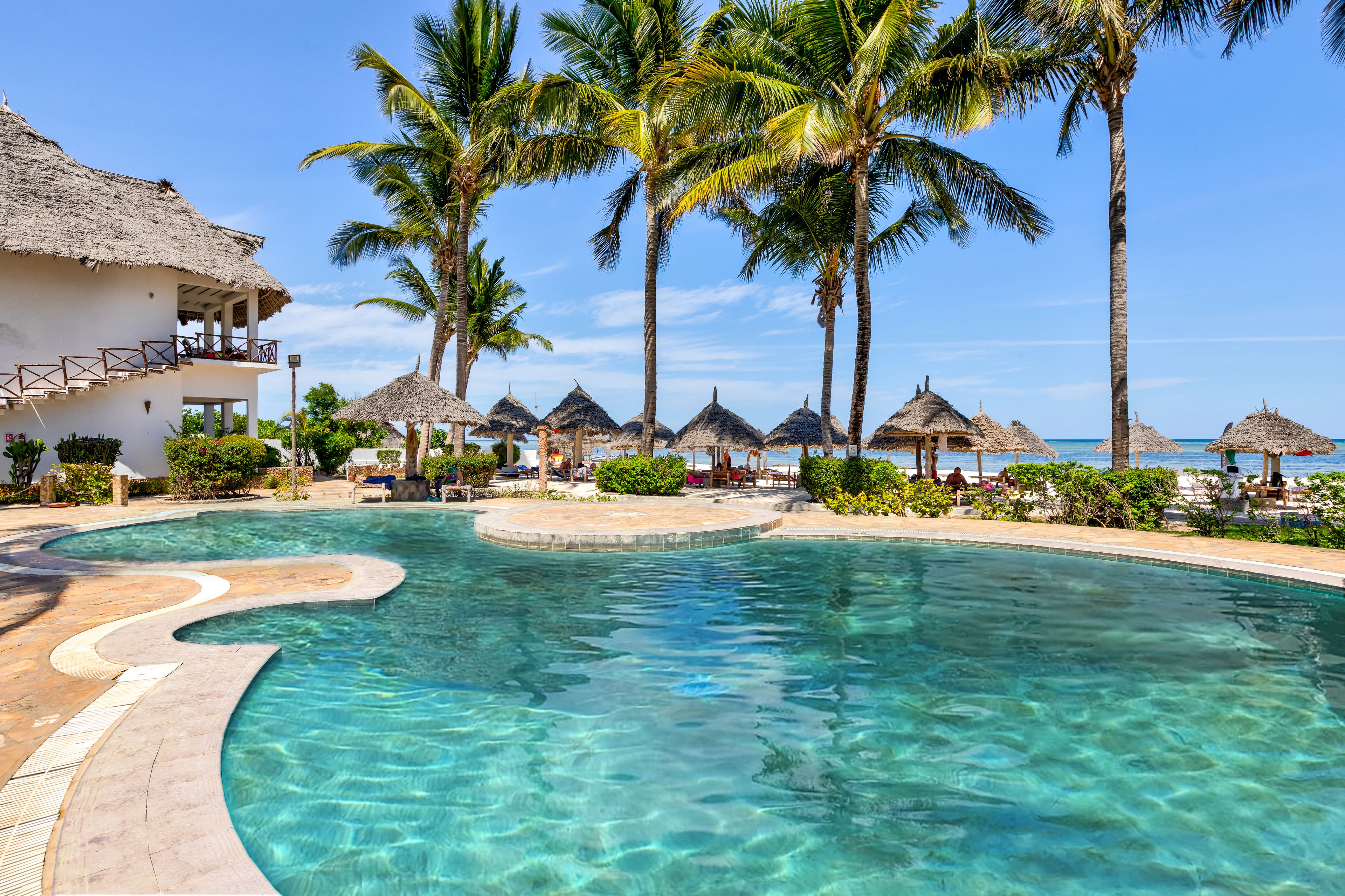 A resort pool with clear water, surrounded by palm trees and thatched umbrellas, with a building featuring a balcony.