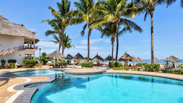 Een zwembad met een golvend ontwerp, omringd door palmbomen en hutten met rieten daken, waarschijnlijk onderdeel van een resort.