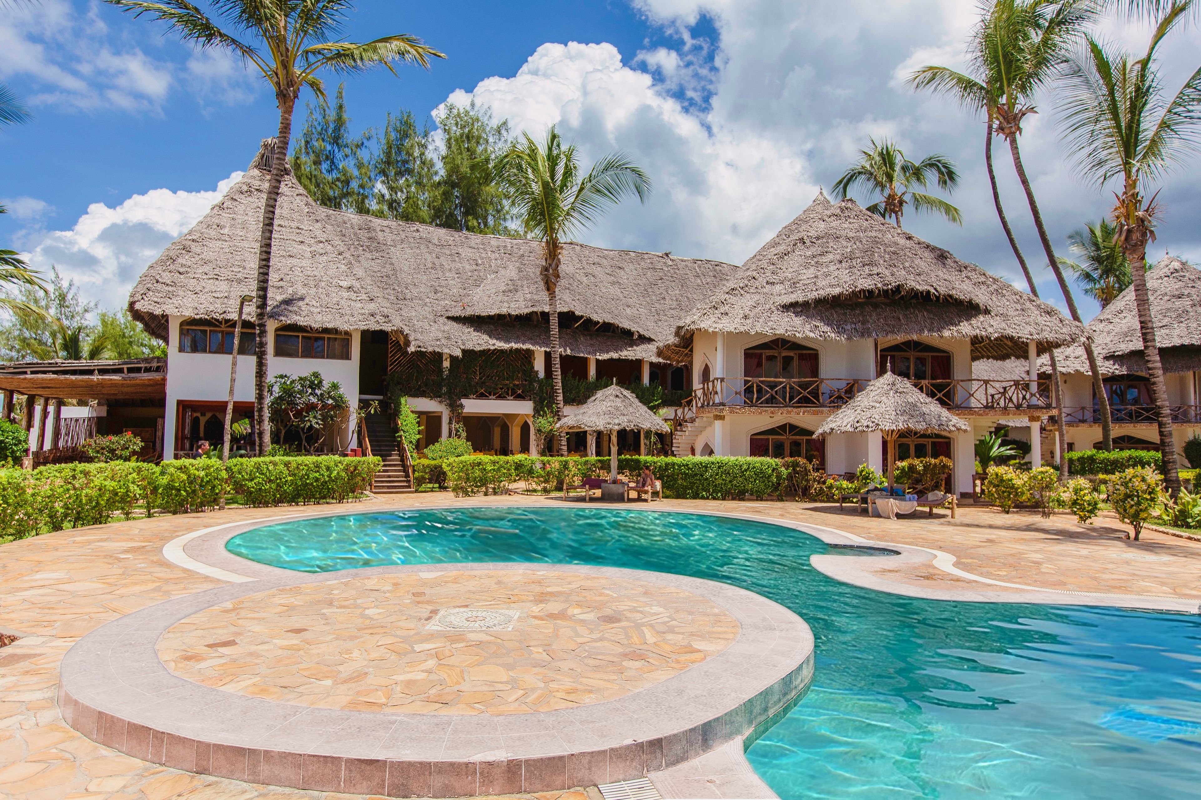 Ein Resort mit strohgedeckten Gebäuden, einem zentral gelegenen Schwimmbad und Palmen.