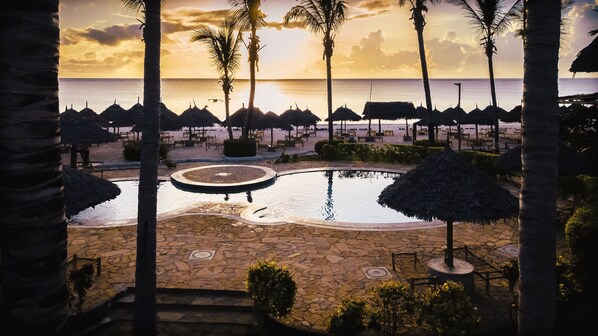 A resort pool area with palm trees, thatched umbrellas, and a view of the ocean at sunset.
