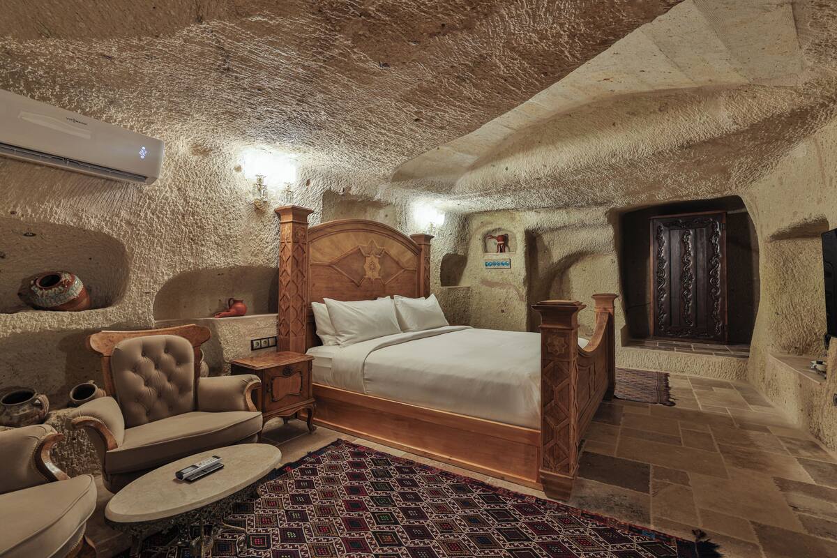 A stone-walled room with a bed, a wooden chair, a small table, and a wall-mounted heater.