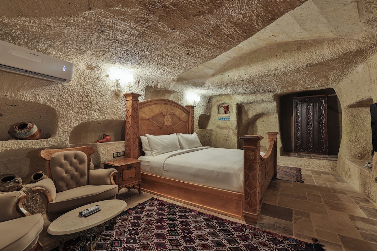 A stone-walled room with a bed, a wooden chair, a small table, and a wall-mounted heater.