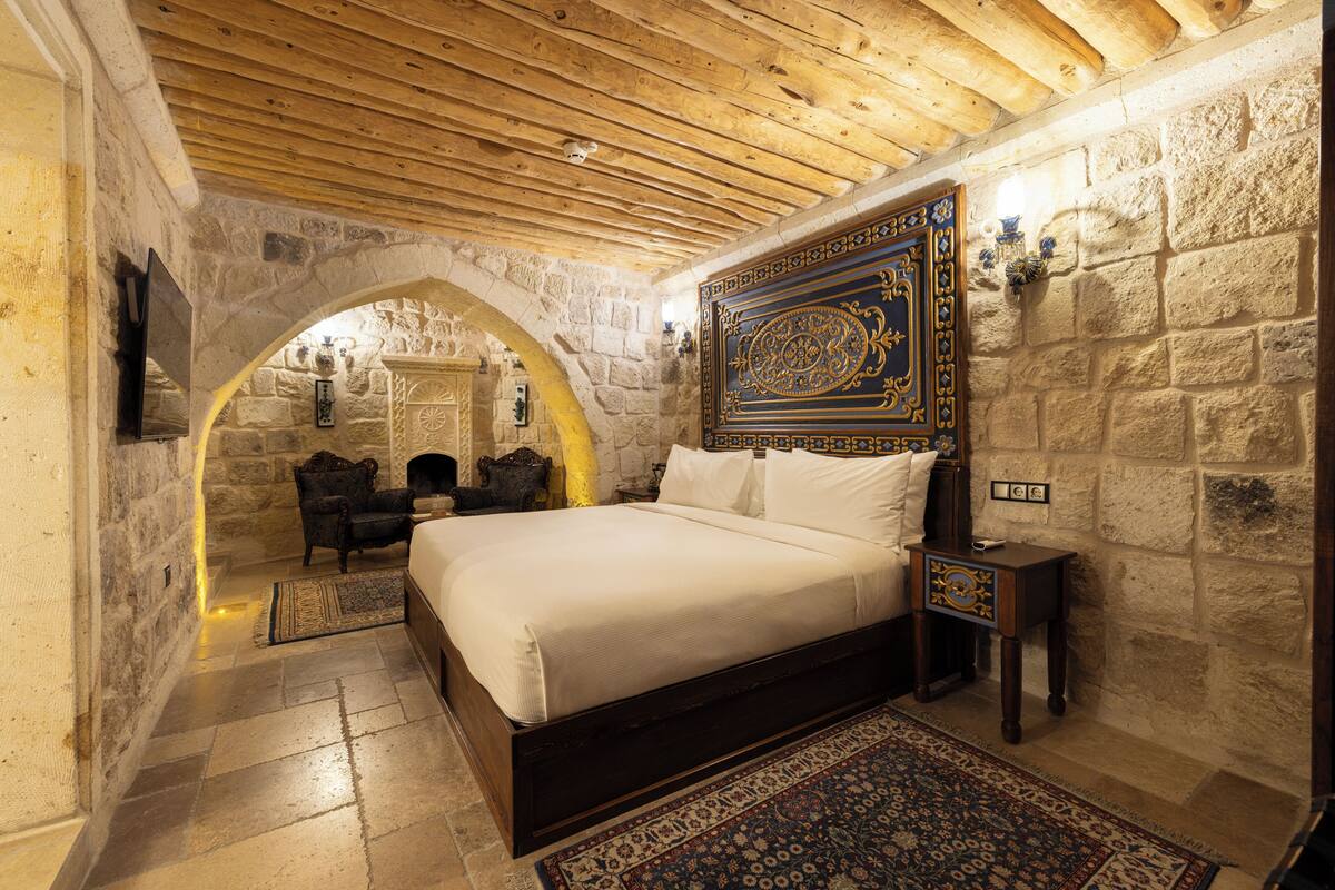 A bedroom with a stone wall, wooden ceiling, a bed with white linens, a patterned rug, and a small table with a lamp.