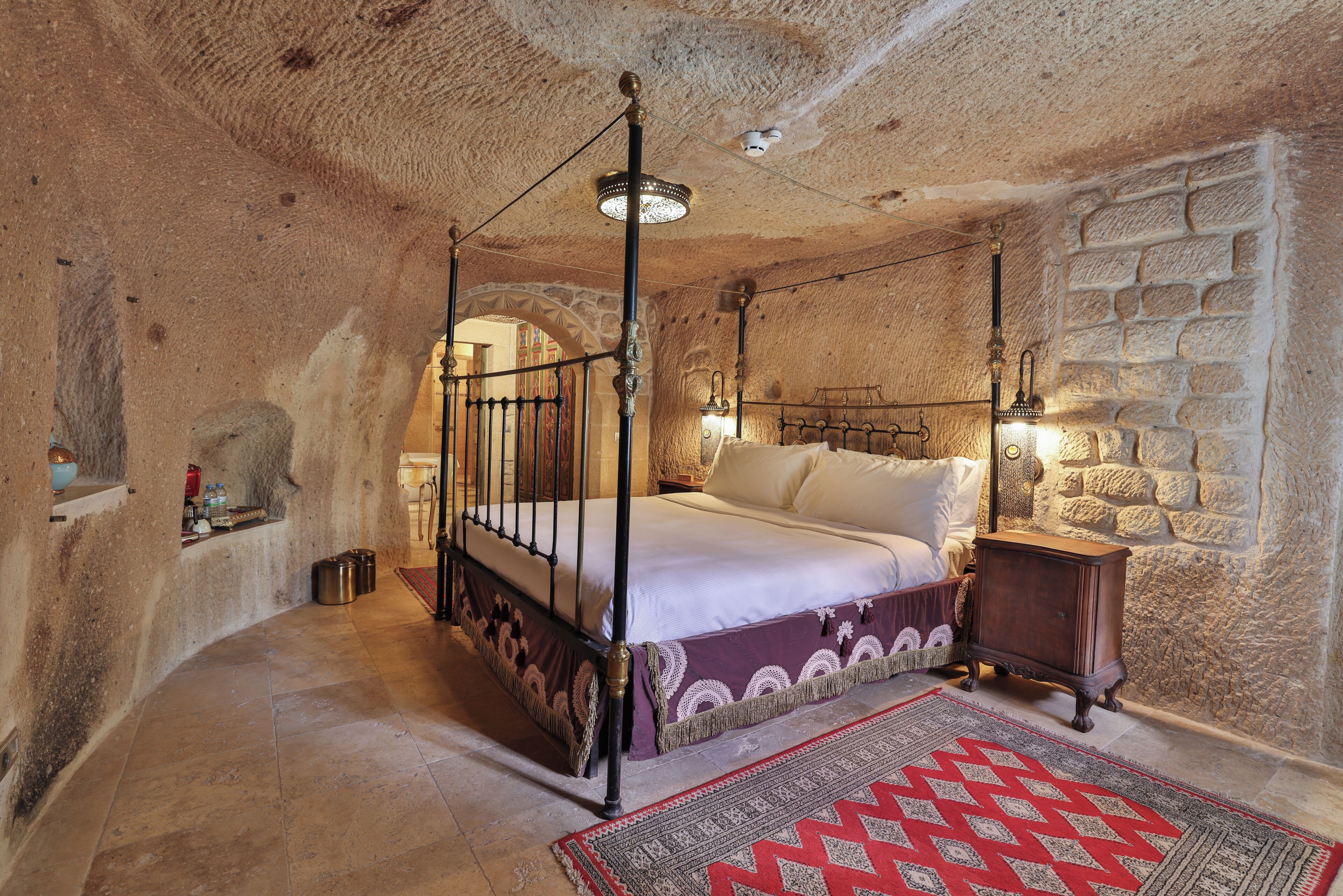 Un lit à baldaquin avec une tête de lit, une table de chevet, un tapis rouge à motifs et des murs en pierre.