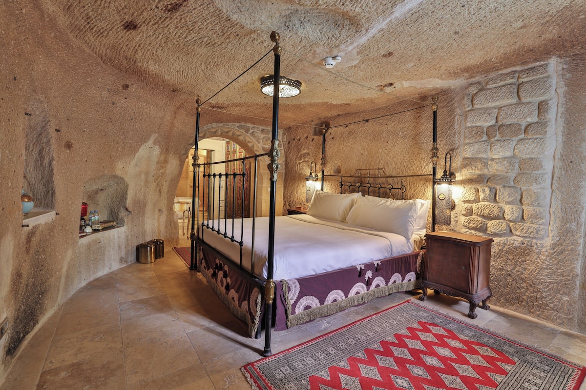 A four-poster bed with a headboard, a nightstand, a red patterned rug, and stone walls.