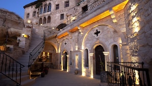 Un edificio histórico de piedra con una escalera que lleva a un balcón, arcos en las entradas y una entrada bien iluminada.
