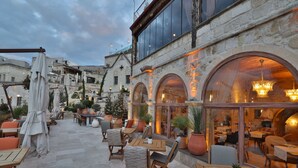 Un café al aire libre con mesas y sillas, plantas en macetas y un edificio de piedra con ventanas de arco.
