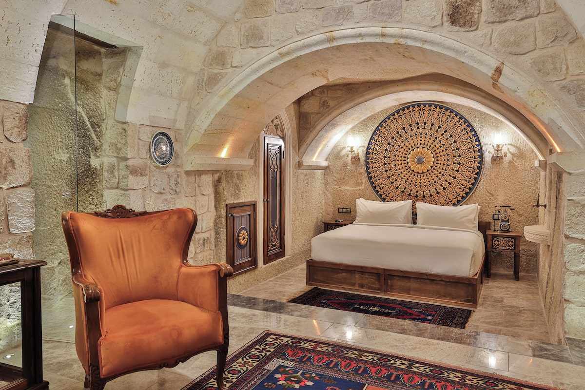 A bedroom with a stone wall, a large bed, a chair, and a decorative ceiling.