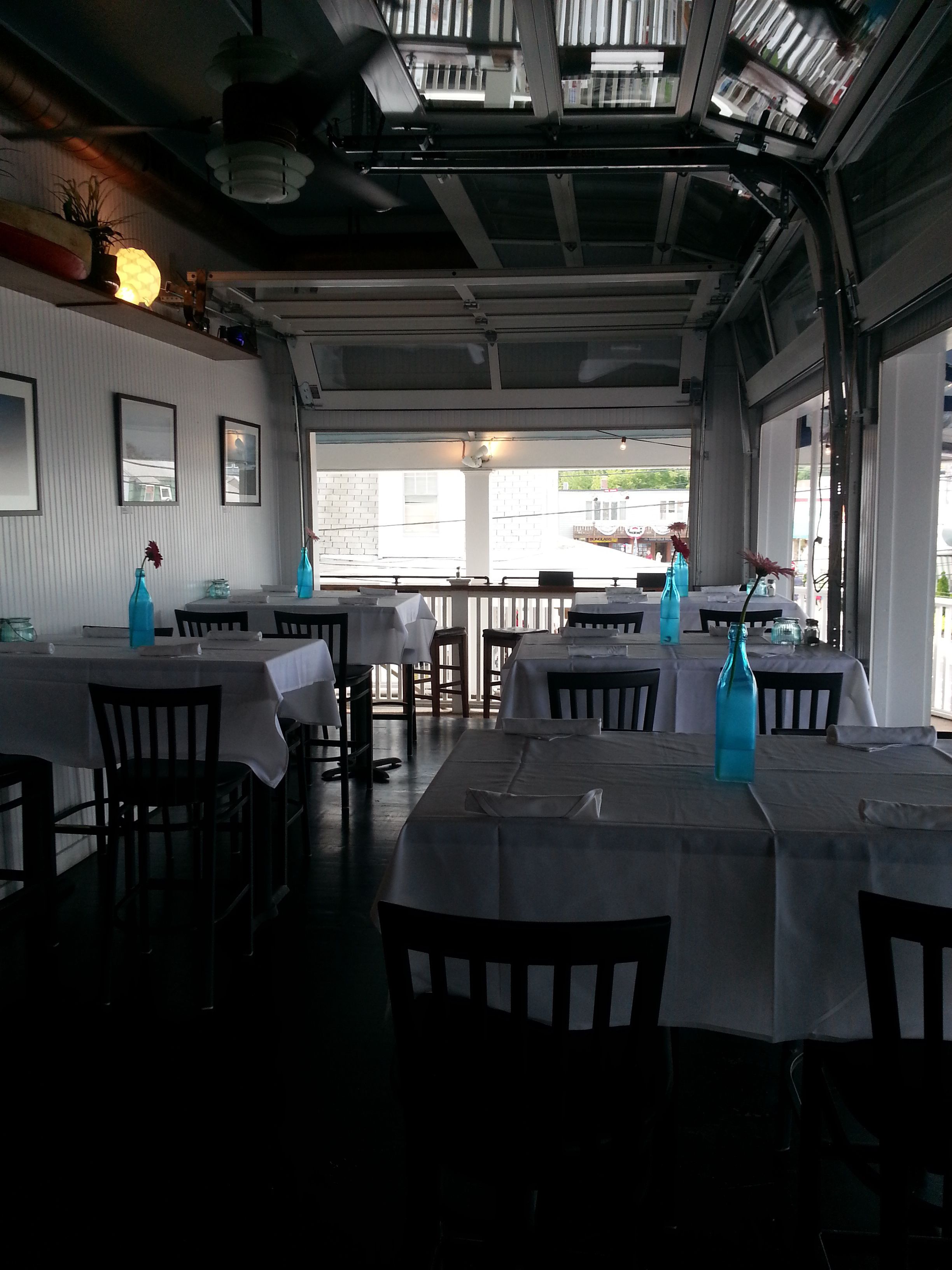 A restaurant with tables set for dining, a view of the street outside, and a ceiling with exposed beams and lighting fixtures.