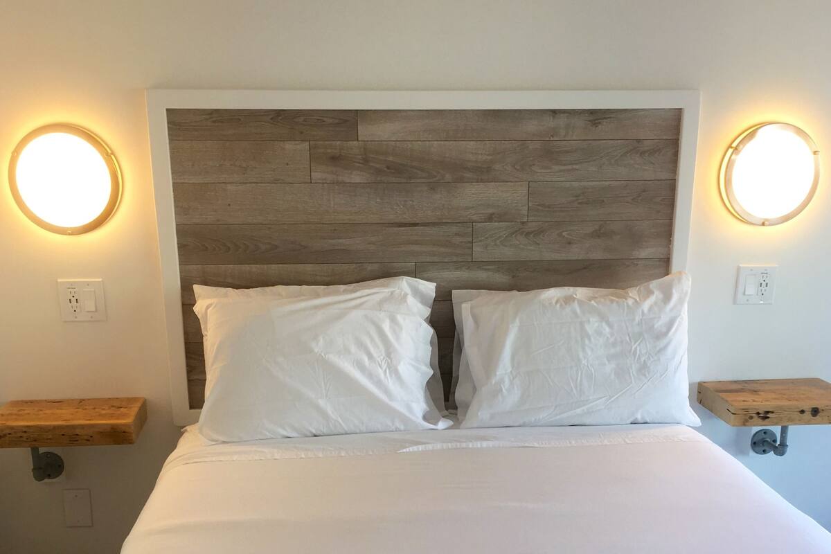 A bed with white linens, a wooden headboard, and two bedside tables with round wall lights.