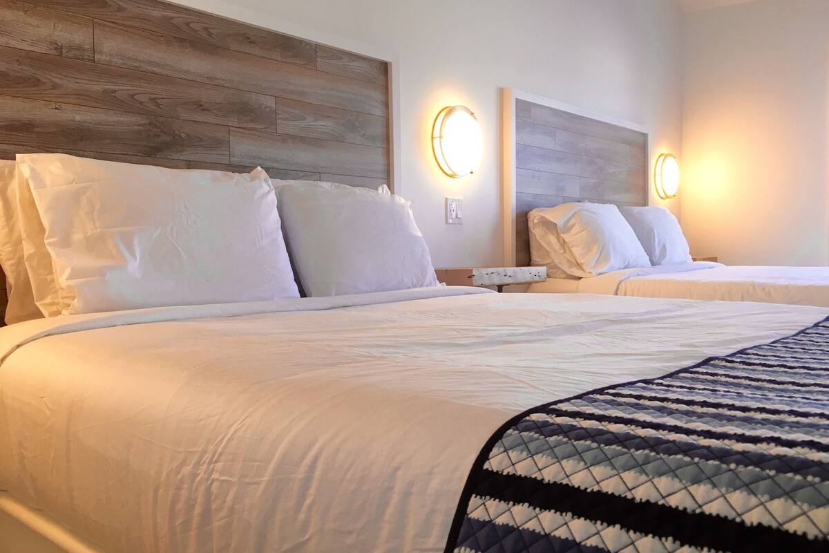 A hotel room with two beds, each with white bedding and headboards. There are wall-mounted light fixtures on either side of the beds.