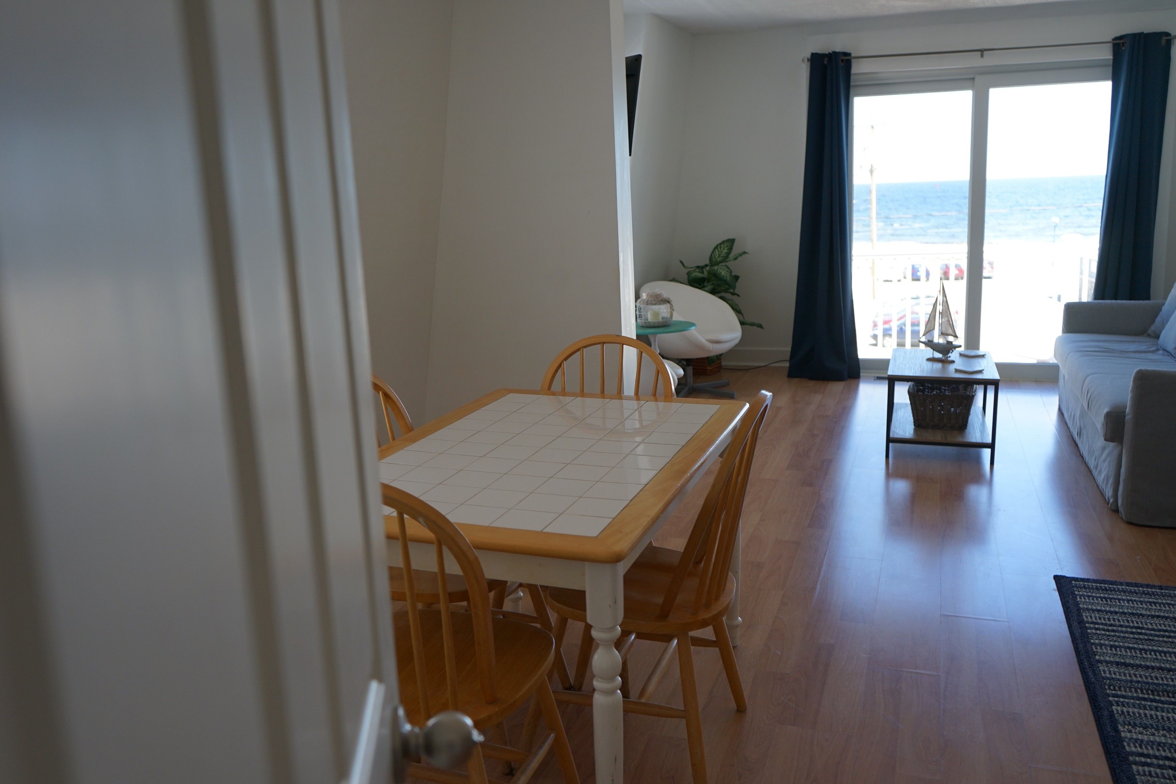 A dining area with a white table and wooden chairs, a small round table with a plant, and a sofa with a view of the sea.