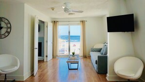 A living room with a ceiling fan, a flat-screen TV, a sofa, a coffee table, and a view of the ocean through a sliding glass door. - Inn on the Blues (York Beach)