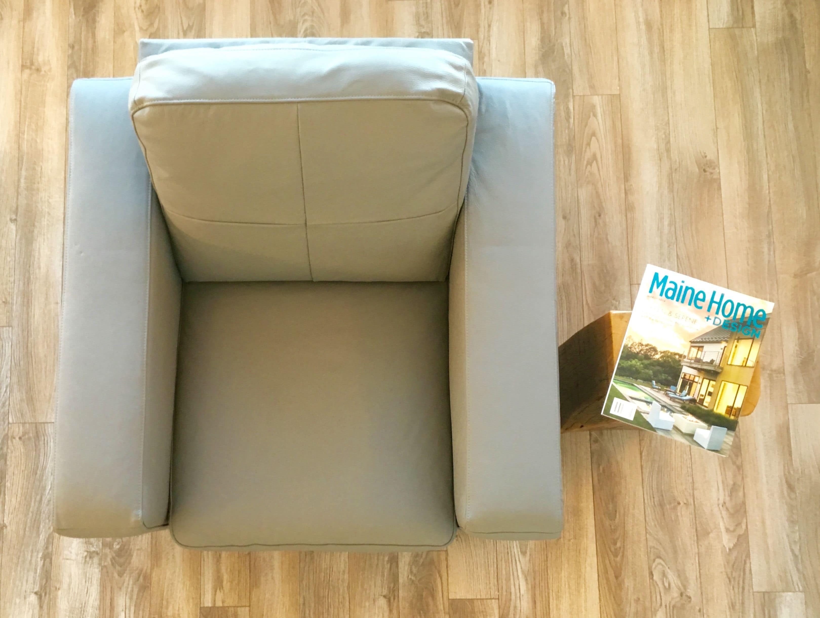 A beige sofa with a cushion, positioned on a wooden floor. A magazine titled 'Maine Home + Design' is placed beside the sofa.