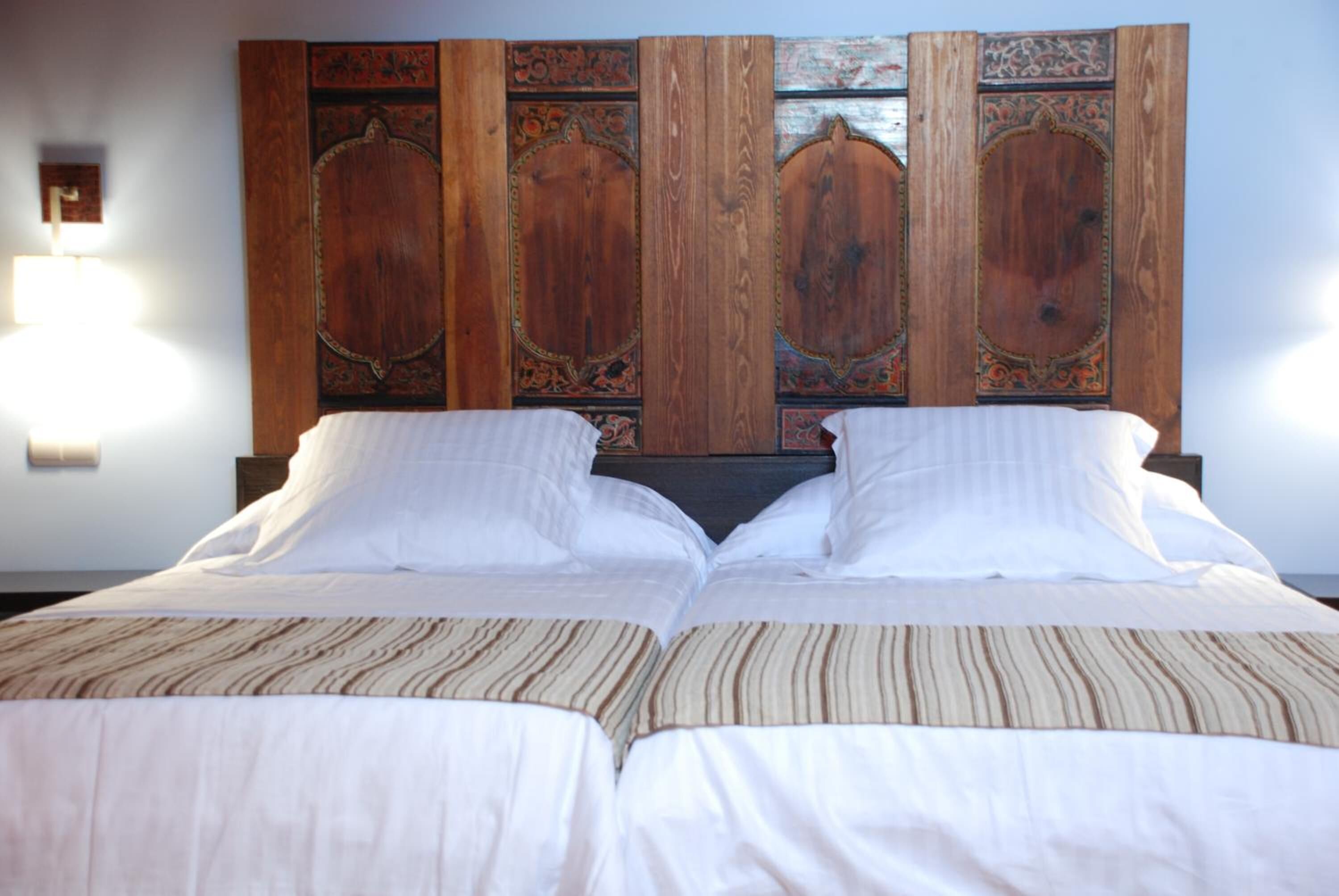 A double bed with a striped bedspread, a wooden headboard with ornate carvings, and a wall-mounted light fixture.