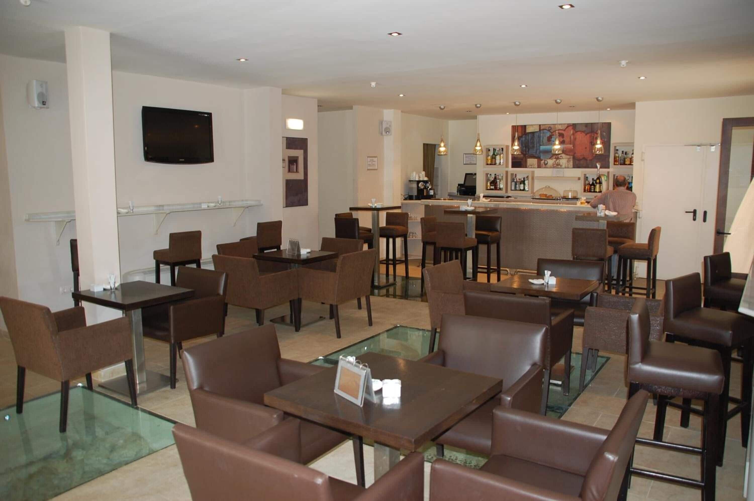 Un hall d’hôtel moderne avec des tables à plateaux de verre et des fauteuils en cuir marron, un bar avec un barman et un téléviseur à écran plat fixé au mur.
