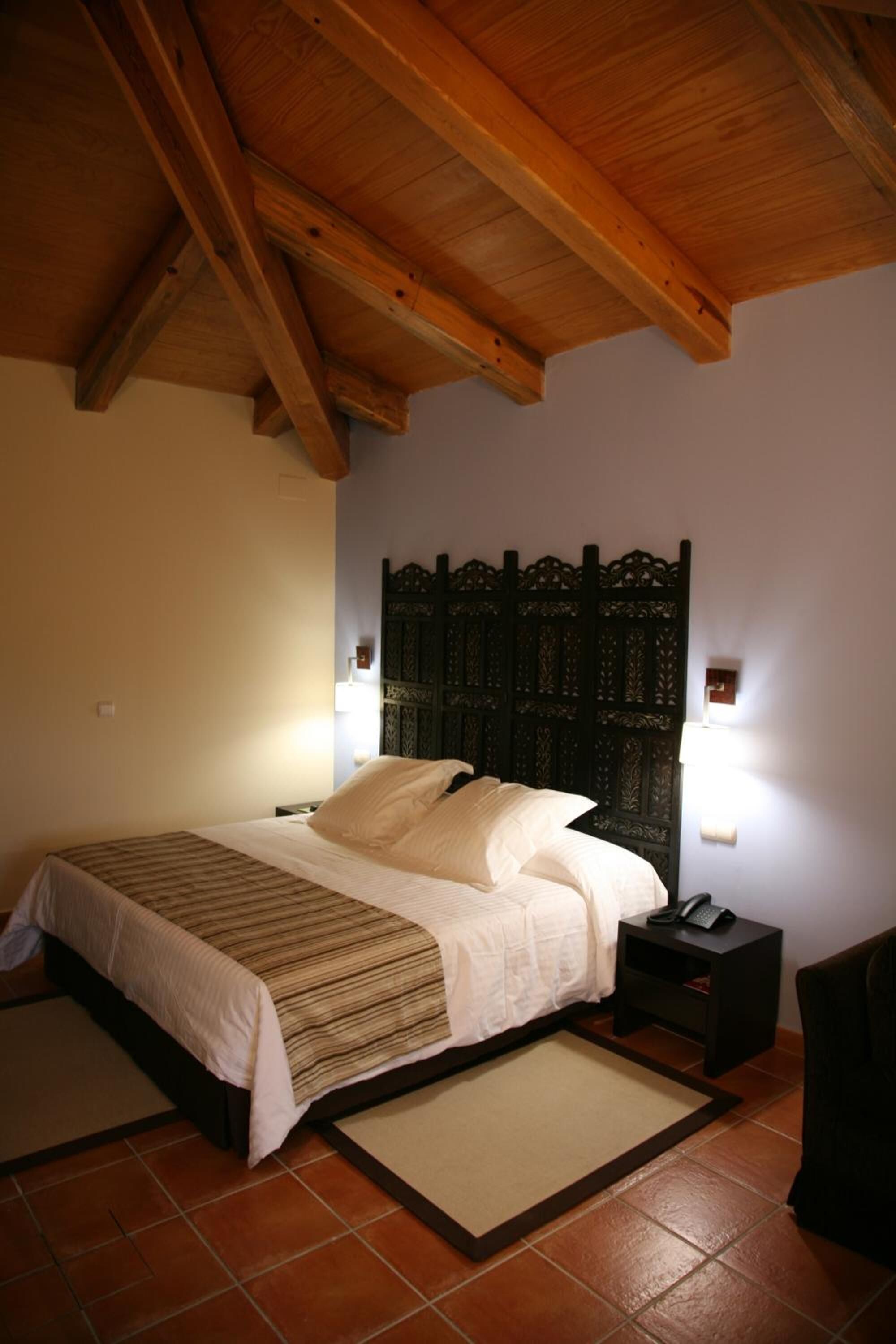 A bedroom with a wooden ceiling, a bed with white bedding, a nightstand with a phone, and a decorative headboard.
