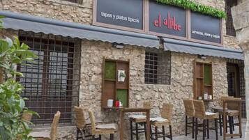 A stone building with a sign reading "el aljibe tapas y platos típicos." There are outdoor tables and wicker chairs.
