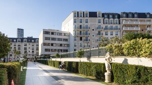 A paved walkway lined with hedges and statues, leading to multi-story residential buildings with balconies.