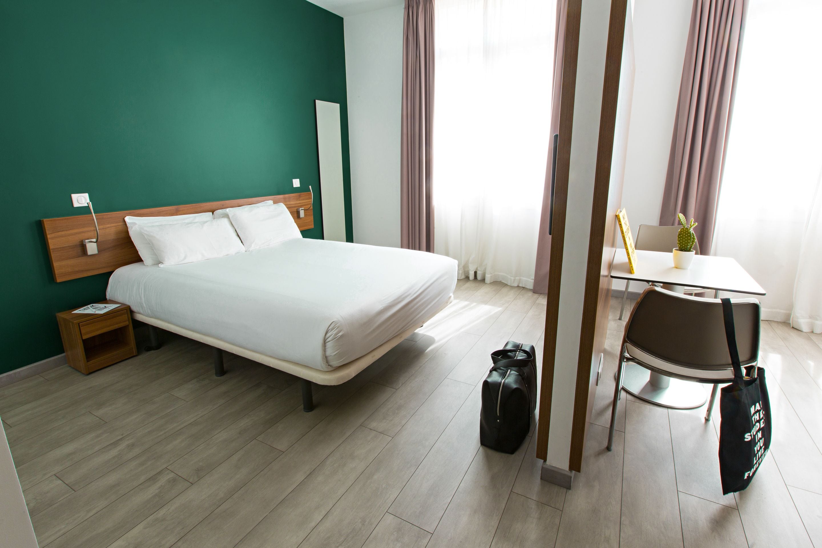A hotel room with a bed, bedside table, chair, and a small table with a plant.