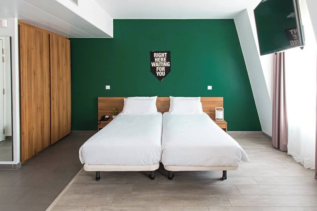 Une chambre avec deux lits, une armoire en bois, un mur d’accent vert, un téléviseur fixé au mur et une pancarte sur laquelle on peut lire :  « C’EST ICI QUE NOUS VOUS ATTENDONS ».