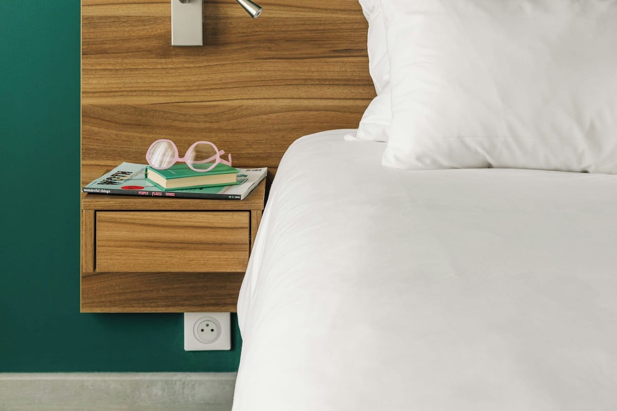 Un lit bien fait, avec des draps blancs, une tête de lit en bois, une table de chevet sur laquelle se trouvent un livre et des lunettes, et une prise électrique.
