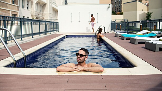 A pool area with a man relaxing in the water, another person walking in the background, and lounge chairs along the poolside.