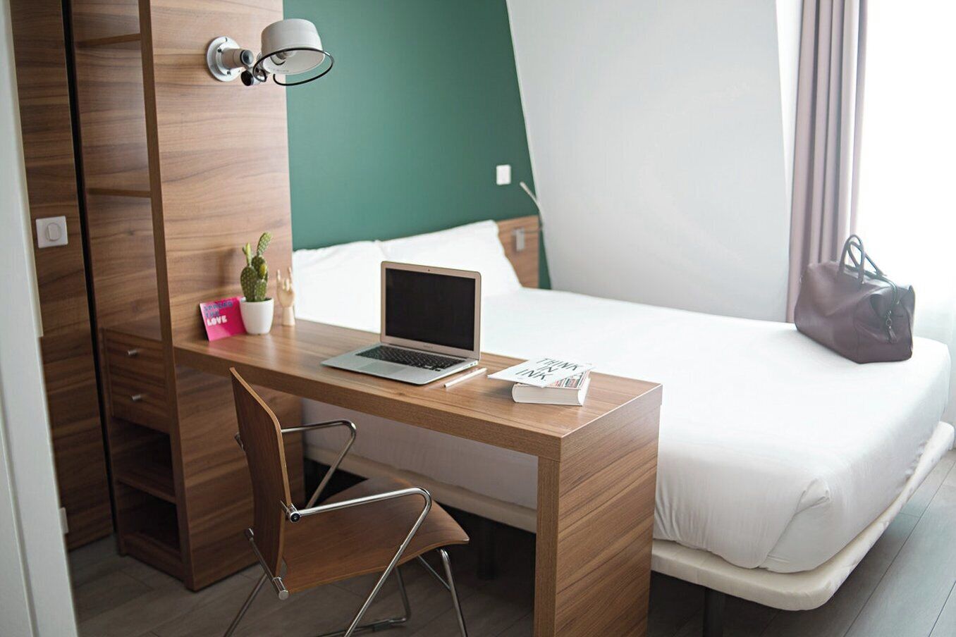 A bedroom with a wooden desk, a laptop, a chair, a bed, a nightstand, a lamp, a suitcase, and a potted plant.