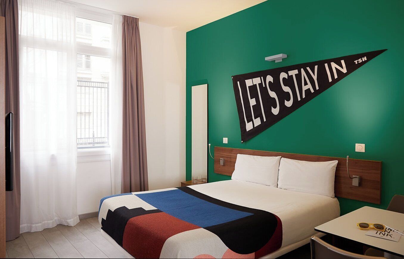 A hotel room with a bed, a nightstand, a table with glasses, and a 'LET'S STAY IN' sign on the wall.