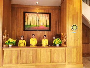 A reception desk with three staff members in yellow uniforms, wooden paneling, a framed painting of a forest, a clock, and a sign reading "MYANMAR". - Queen's Park Hotel (Yangon)